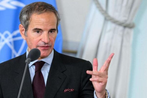 Rafael Grossi, gesturing with his hand and with a serious expression, speaking into a microphone. The blue IAEA flag hangs behind him. He has short grey hair and is wearing a black suit with blue striped shirt and maroon tie.