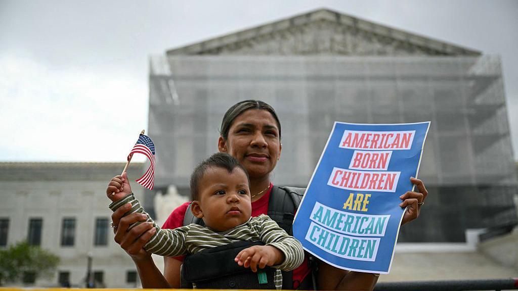 A mother with her infant son hold a US flag and sign saying