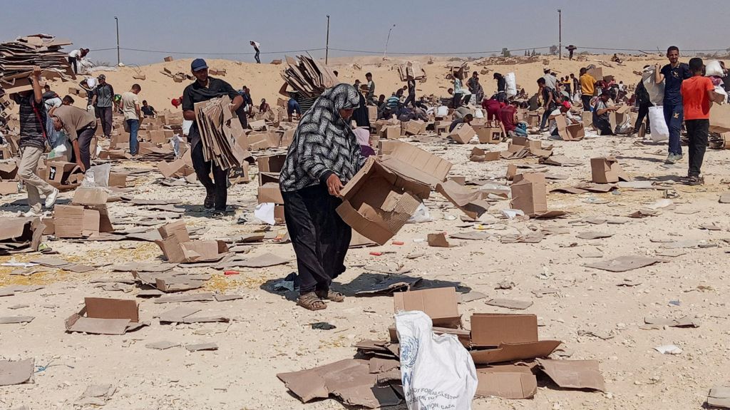 Palestinians gather to collect what remains of relief supplies from the distribution center of the U.S.-backed Gaza Humanitarian Foundation, in Rafah, in the southern Gaza Strip. Empty cardboard boxes litter the arid ground.